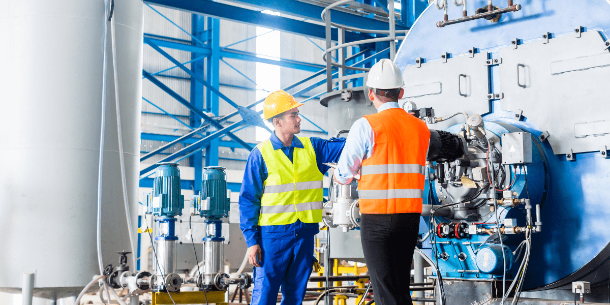 A factory worker in a yellow hard hat talks with a manager in an orange vest while inspecting a large blue industrial boiler in a plant.