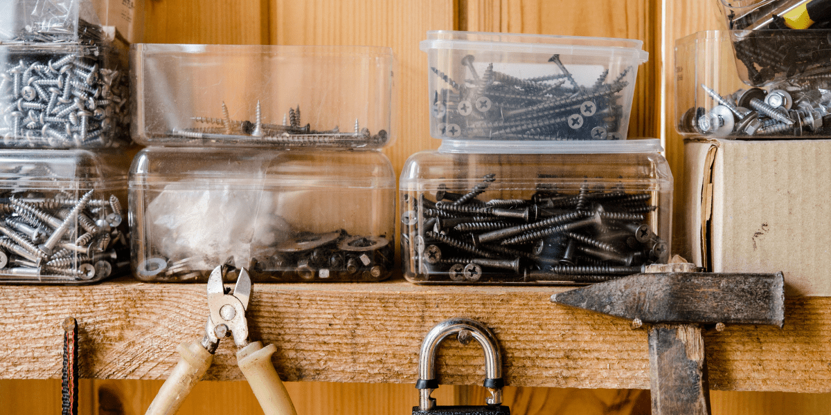 A wooden workshop shelf with clear plastic bins of screws. Below the shelf, pliers, a padlock, and a hammer are stored.