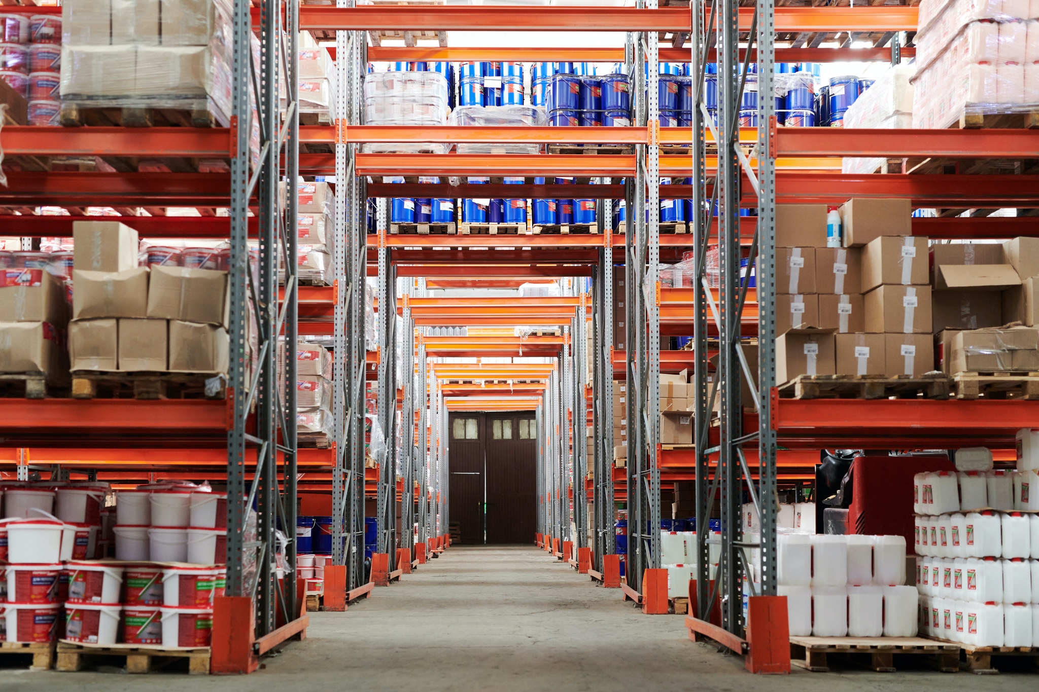 A view down a warehouse aisle lined with tall orange racking. The shelves are fully stocked with boxes, barrels, and other inventory.