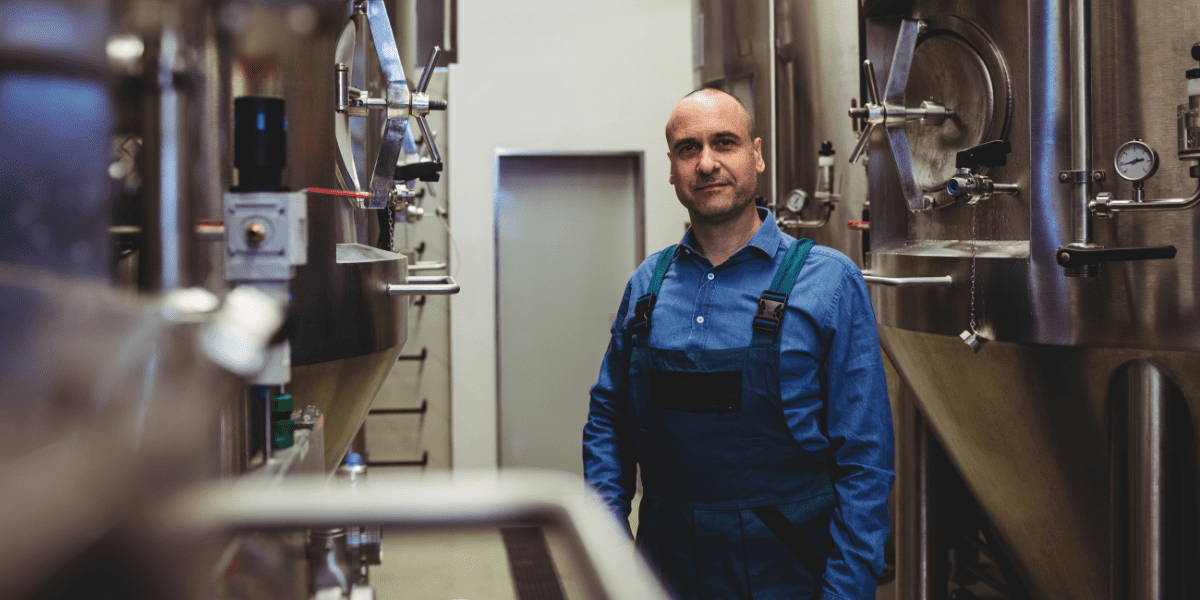 A male worker in blue overalls stands between large, stainless steel fermentation tanks in a brewery or food processing plant.