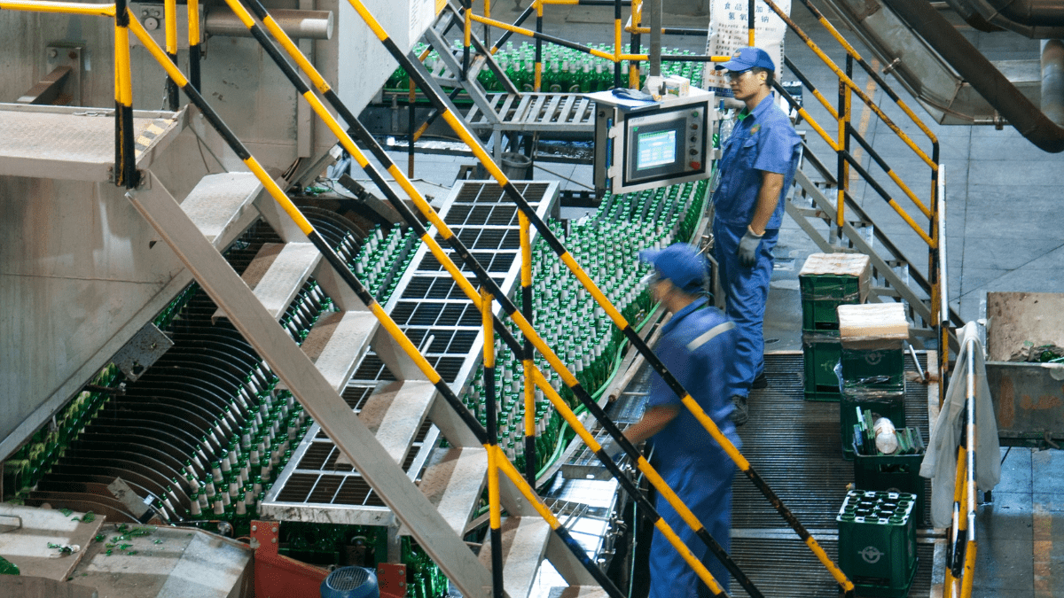 High-angle view of two workers in a bottling plant, one at a control panel, managing a production line of green glass bottles on a conveyor belt.