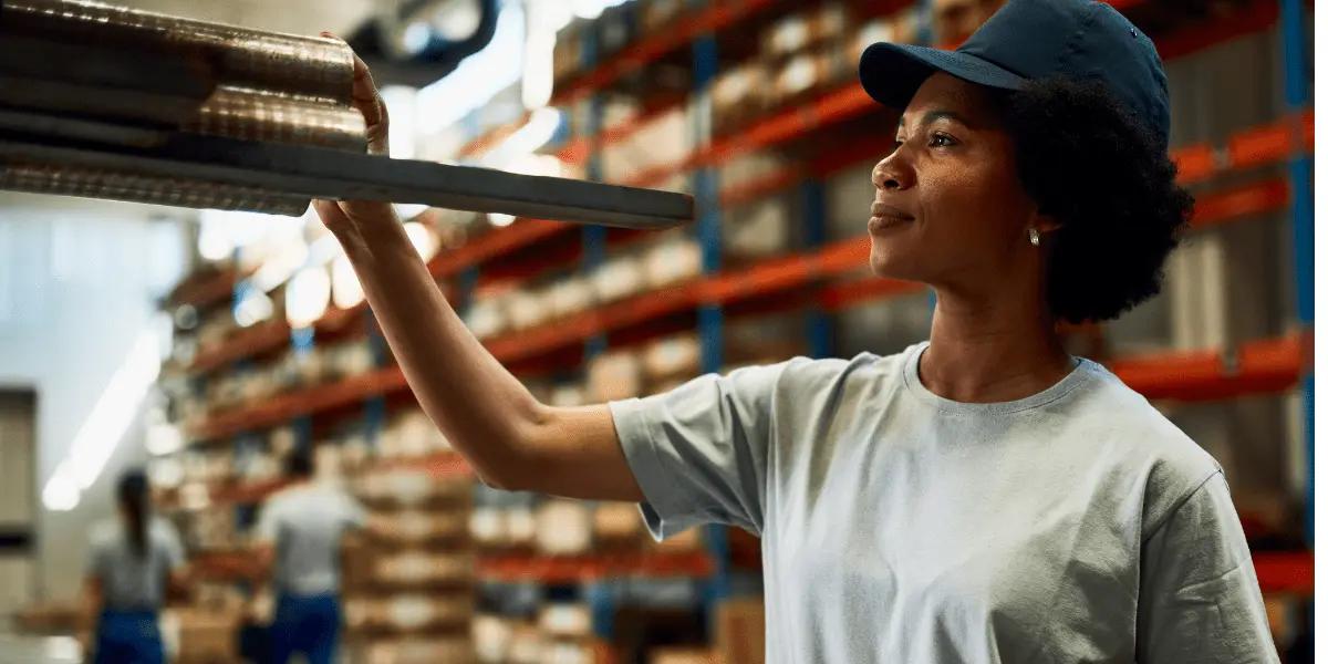 A warehouse worker in a cap and t-shirt reaches for supplies on a shelf.