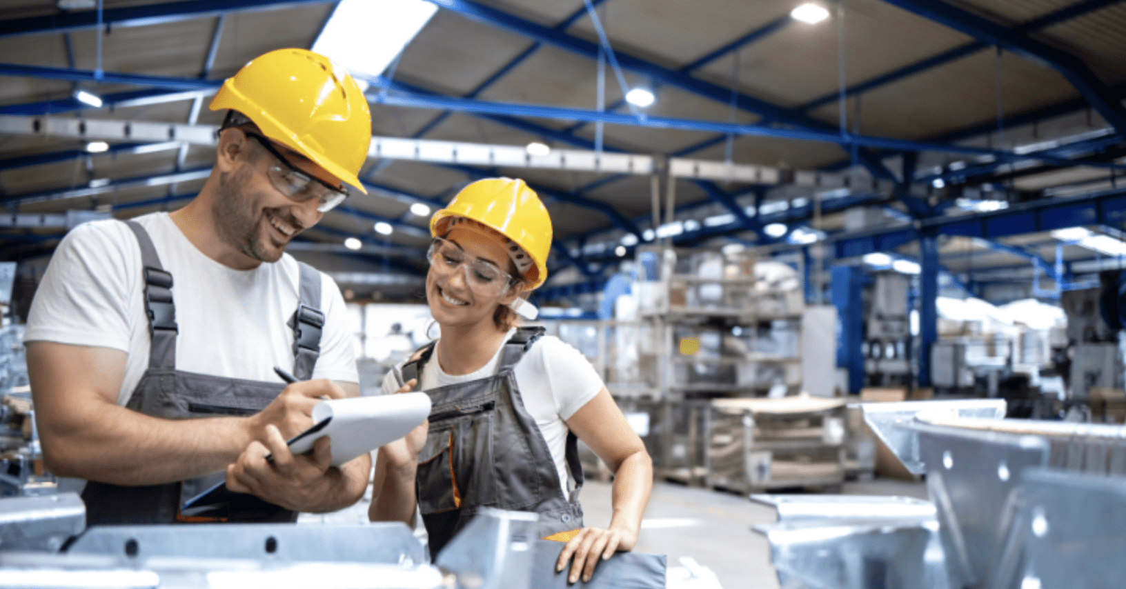 Two smiling factory workers in yellow hard hats and safety goggles collaborate over a clipboard on a busy manufacturing plant floor.