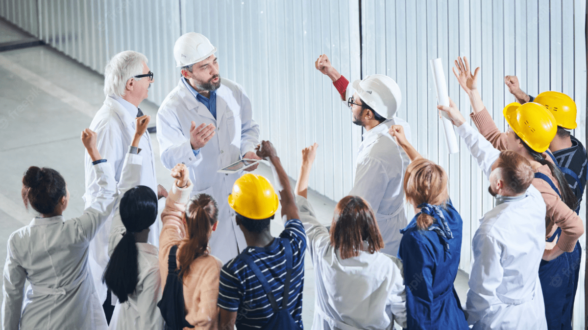 A manager in a hard hat and lab coat leads a team huddle. The diverse group of workers has their hands raised.