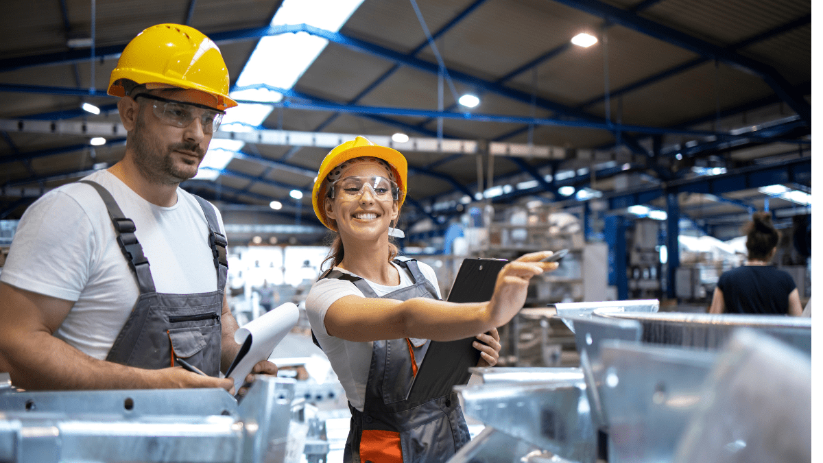 A smiling female worker with a clipboard points something out to her male co-worker during an inspection on a busy factory floor.