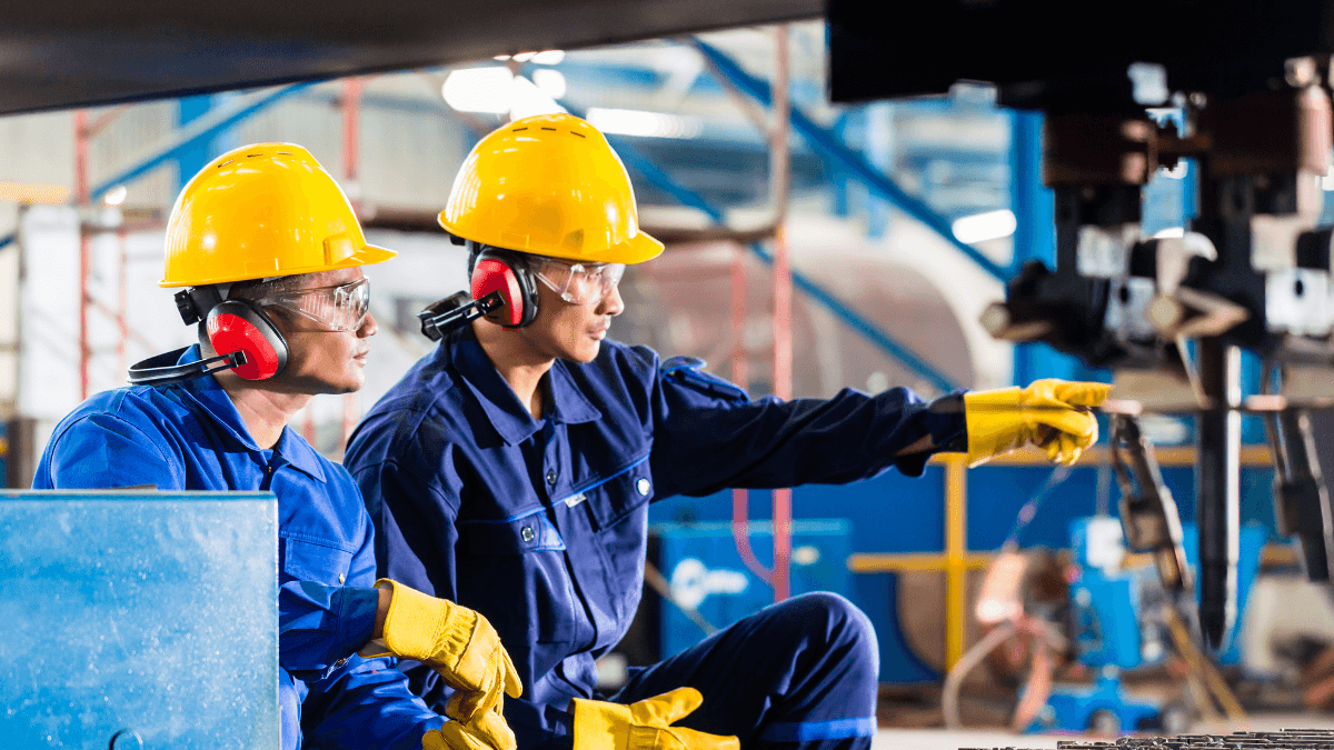 Two factory workers in hard hats and earmuffs inspect industrial machinery, with one worker pointing to a component during the check.