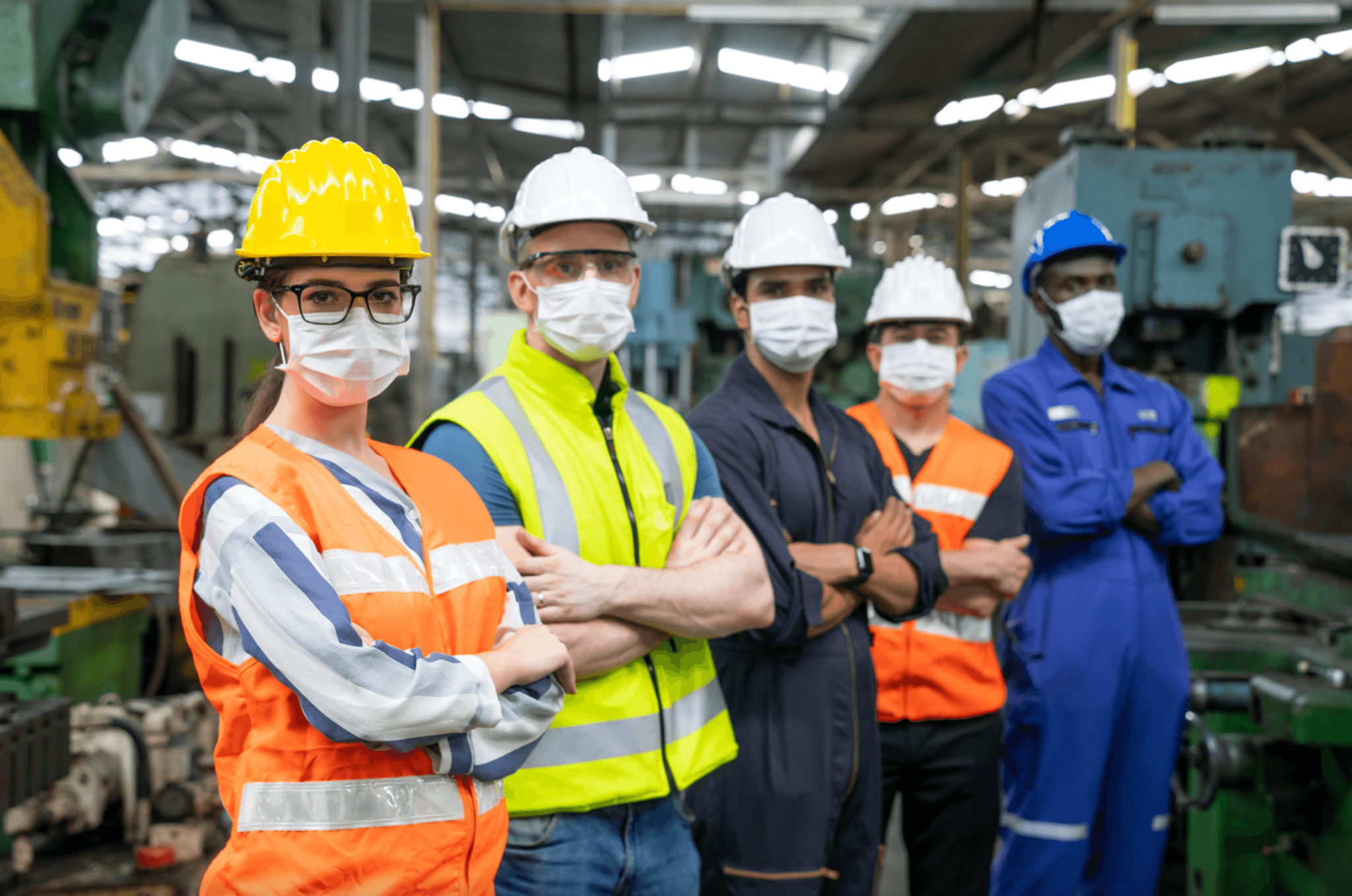 A diverse team of five factory workers in hard hats, face masks, and safety vests stand in a line with arms crossed on a manufacturing plant floor.