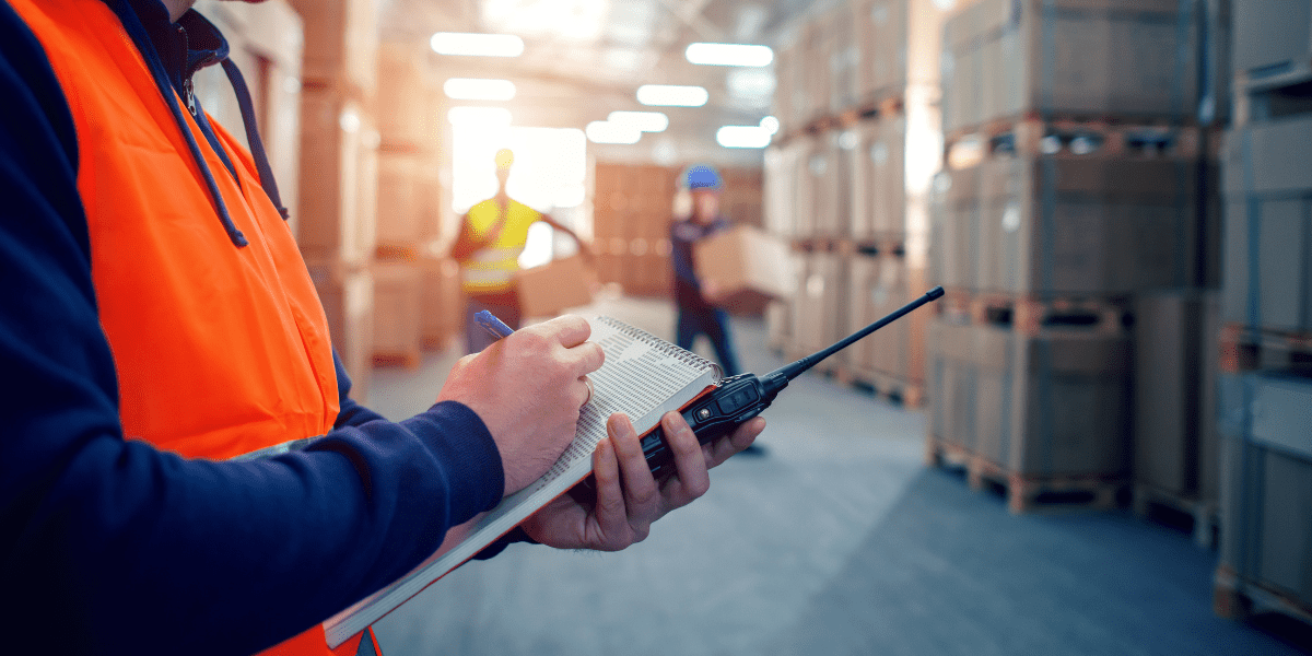 A close-up of a warehouse manager in an orange vest writing on a notepad and holding a walkie-talkie, with other workers moving boxes in the background.