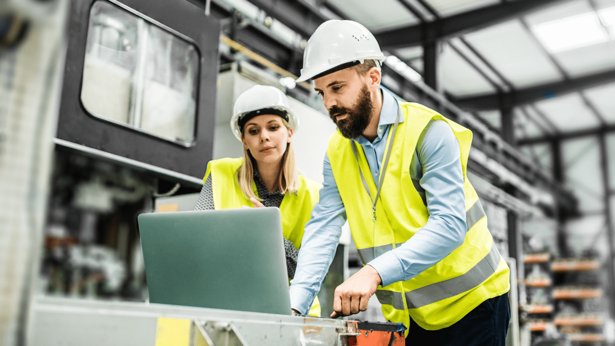 Two industrial engineers, a man and a woman in hard hats and vests, review information on a laptop in a factory setting.