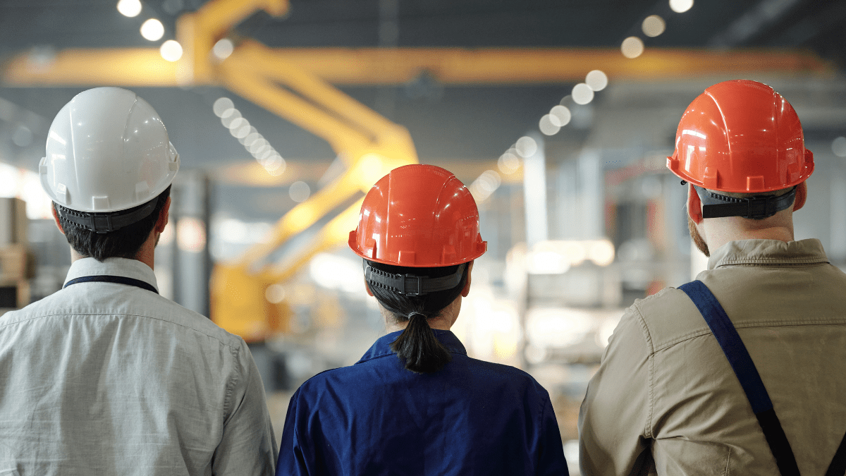 The back of three workers in white and orange hard hats, a woman and two men, observing a large factory floor with an overhead crane.