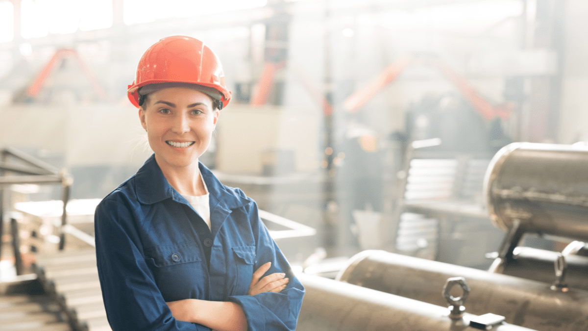 A smiling female industrial worker in a blue coverall and an orange hard hat stands with her arms crossed in a brightly lit factory.