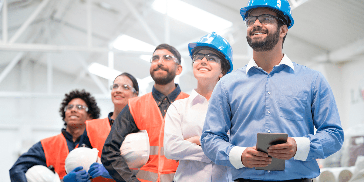 A diverse team of five industrial workers and managers, some in hard hats and vests, smile and look up confidently in a factory setting.