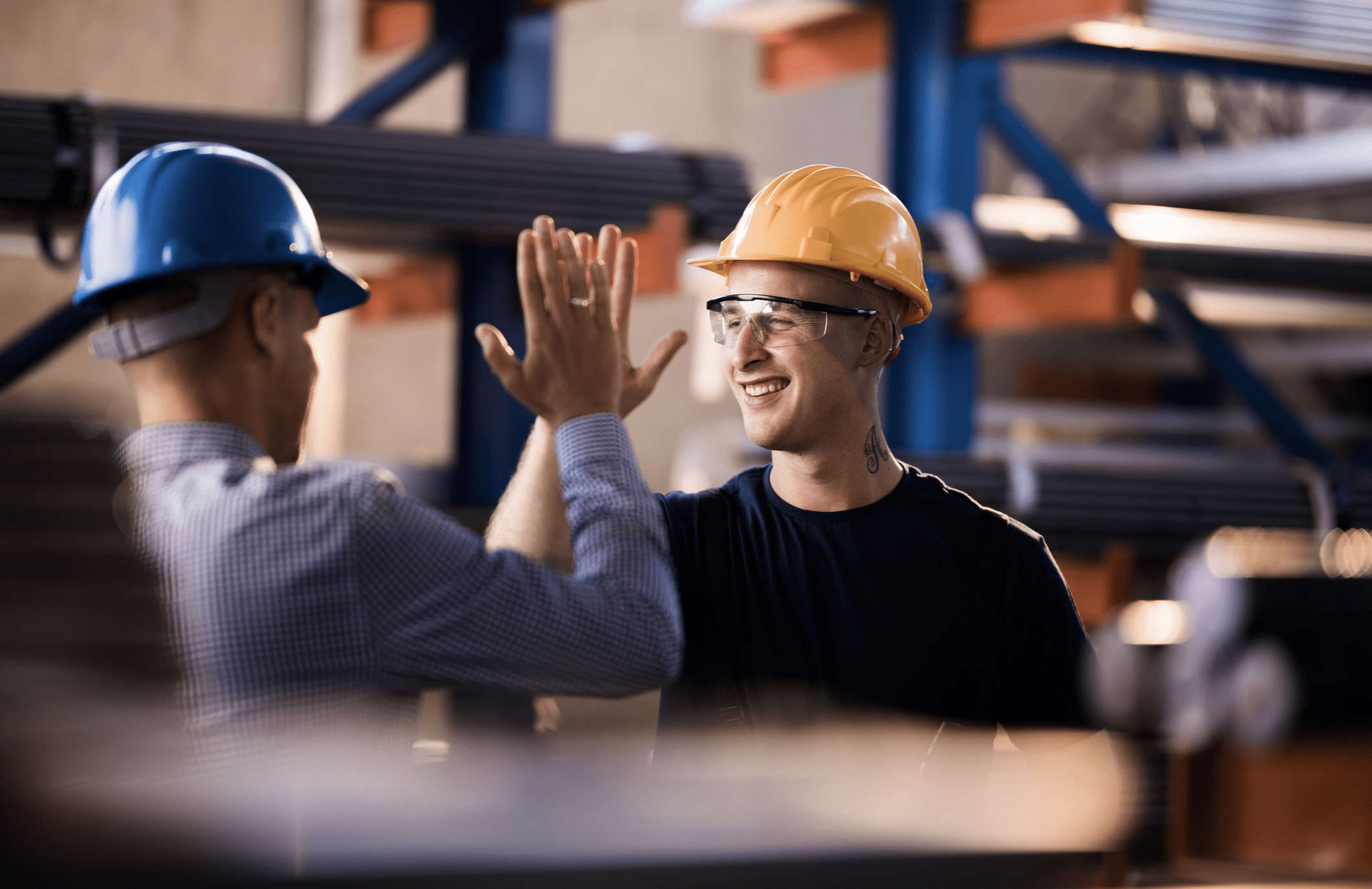 Two smiling industrial workers in hard hats give each other a high-five, celebrating a success or achievement on the factory floor.