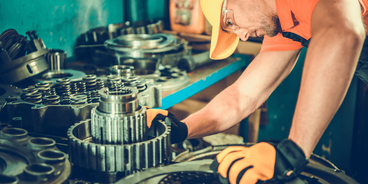 A mechanic in a yellow hard hat and safety glasses carefully inspects a large gear assembly on a workbench full of machine parts.