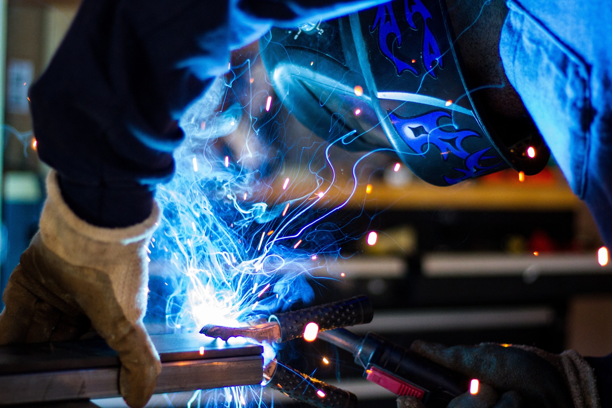 A welder wearing a protective mask and glove joins two pieces of metal, creating a brilliant flash of blue light and hot sparks in a workshop.