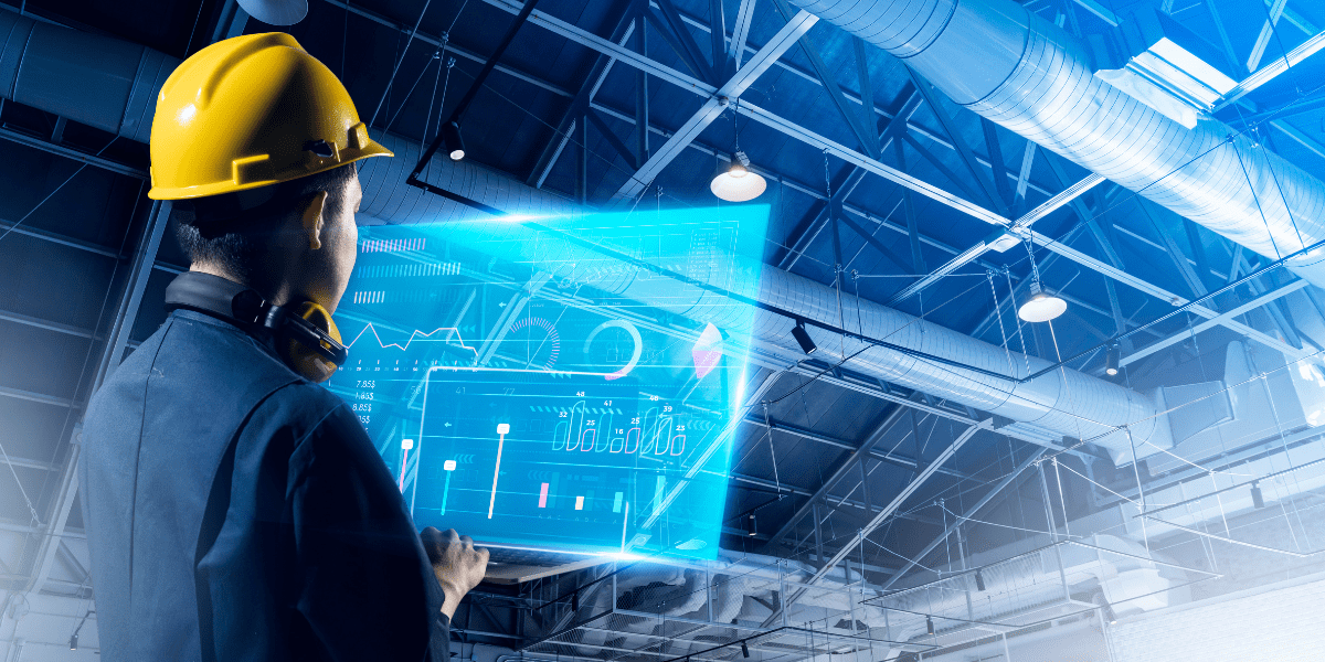 An engineer in a yellow hard hat stands in a factory, viewing a glowing blue holographic data dashboard on a transparent tablet.