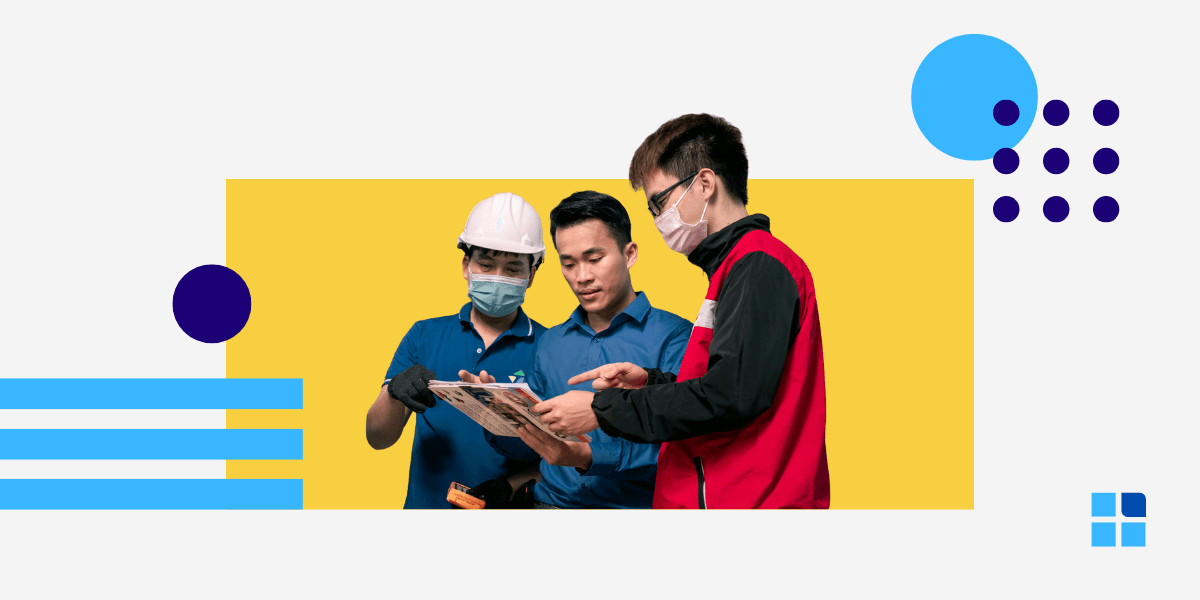Three factory workers in hard hats and face masks collaborate over a document.