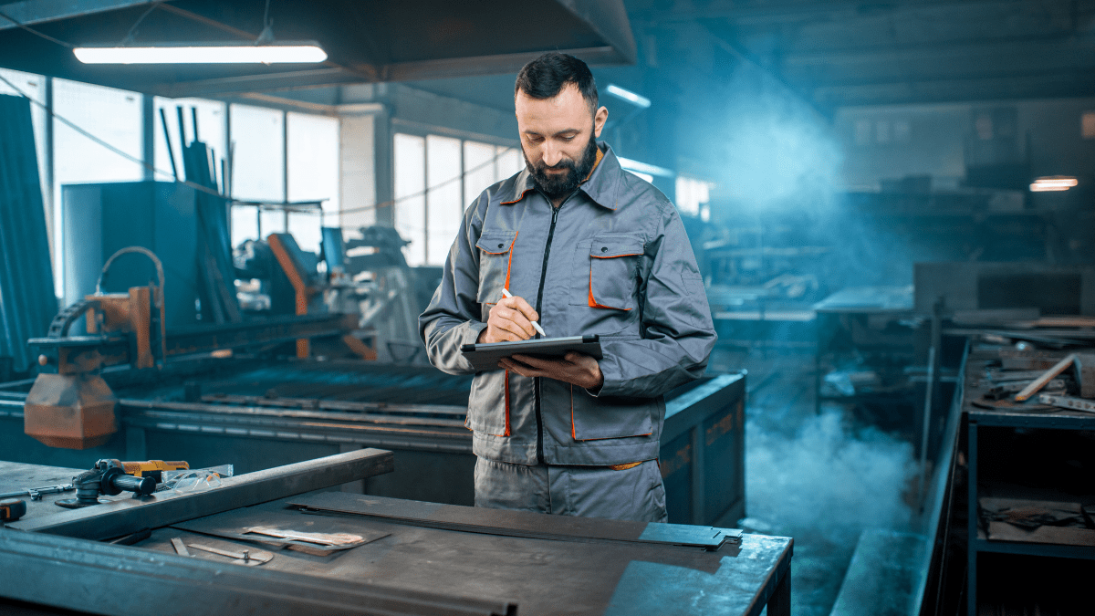 An industrial worker in a grey uniform uses a tablet with a stylus, standing by a workbench in a smoky metal fabrication shop.
