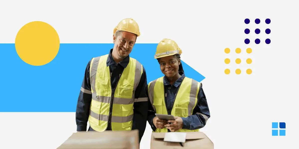 Two construction workers in safety vests and helmets smile while standing at a table with boxes.
